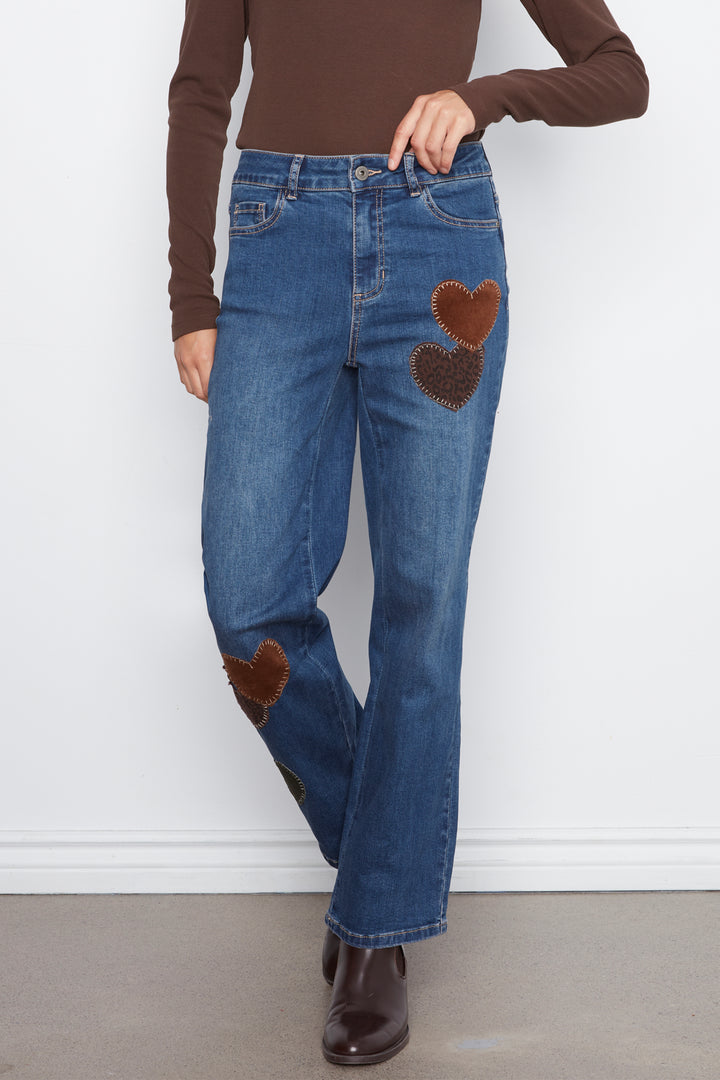A person wearing blue jeans with brown heart-shaped patch details on the thighs and knees, paired with a brown long-sleeve top and dark brown ankle boots, standing against a white wall.