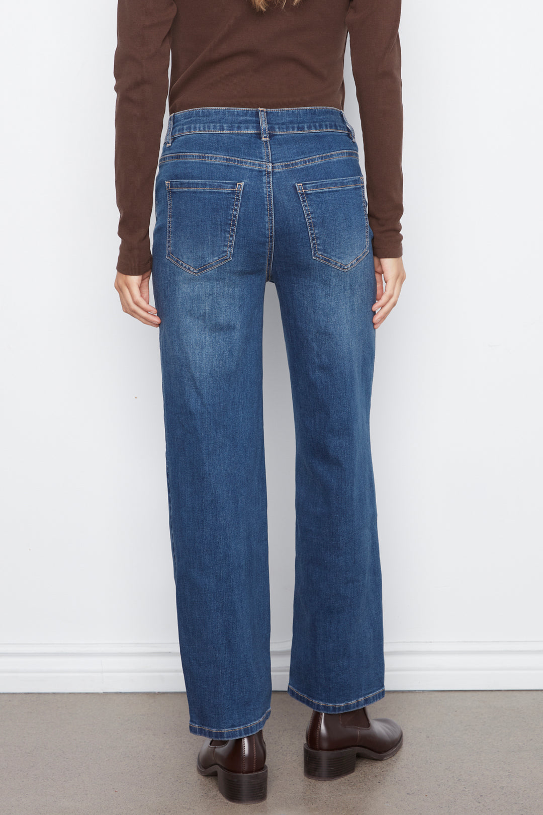 Rear view of a person wearing blue wide-leg jeans, a brown long-sleeve top, and brown leather boots against a plain white wall.