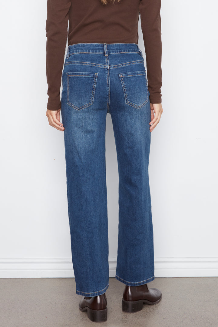 Rear view of a person wearing blue wide-leg jeans, a brown long-sleeve top, and brown leather boots against a plain white wall.