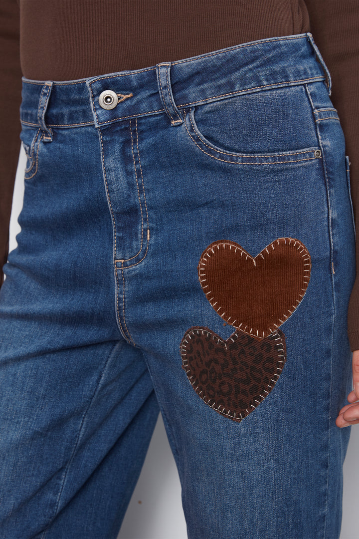 Close-up of blue denim jeans with two brown heart-shaped patch details on the front, worn with a brown ribbed top. A hand is partially visible on the right side.