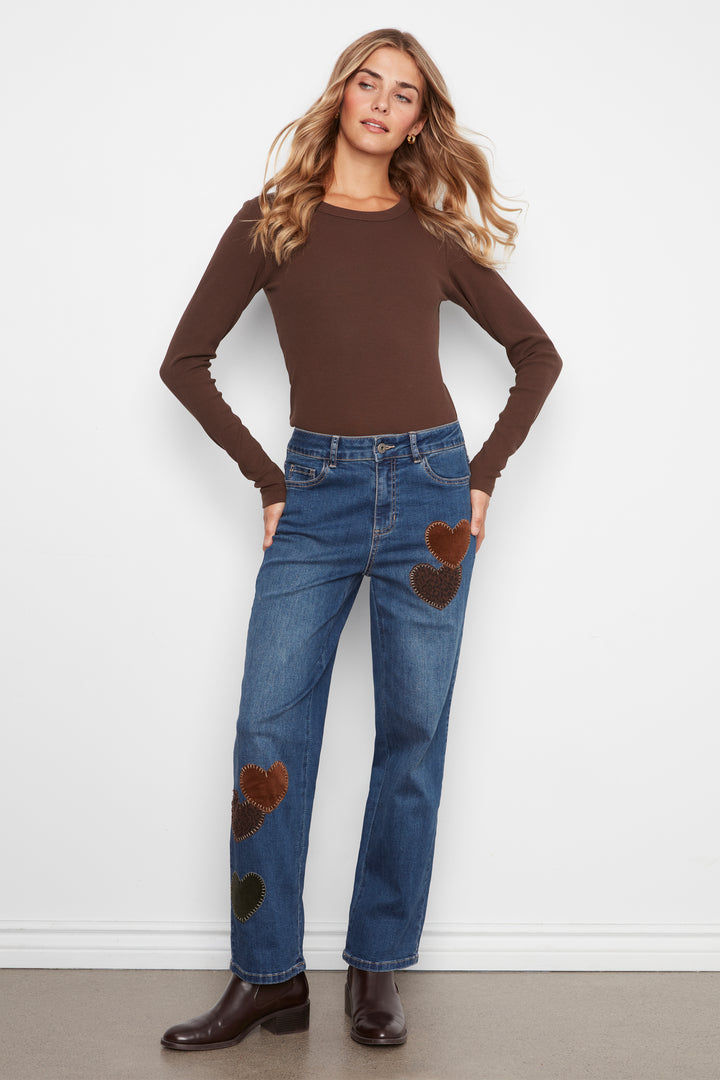 A woman modeling a brown long-sleeve crewneck top and blue jeans with brown heart patches on the thighs and knees, standing against a white wall.