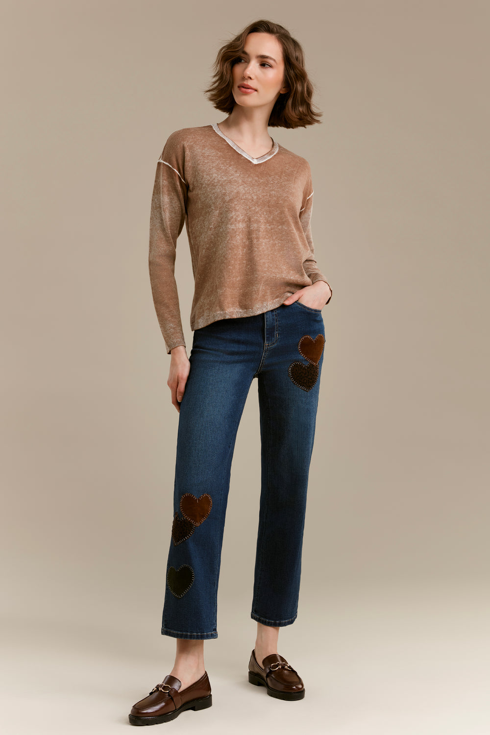 A woman modeling a beige knit long-sleeve sweater and dark blue jeans with brown heart patches, paired with brown leather loafers, against a solid beige studio background.