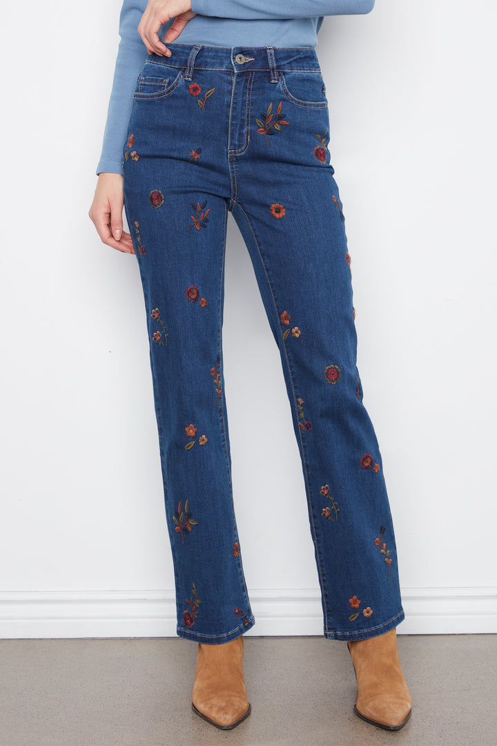 A person wearing blue denim jeans with small embroidered floral details, paired with a light blue long-sleeve top and tan ankle boots, standing against a white wall.