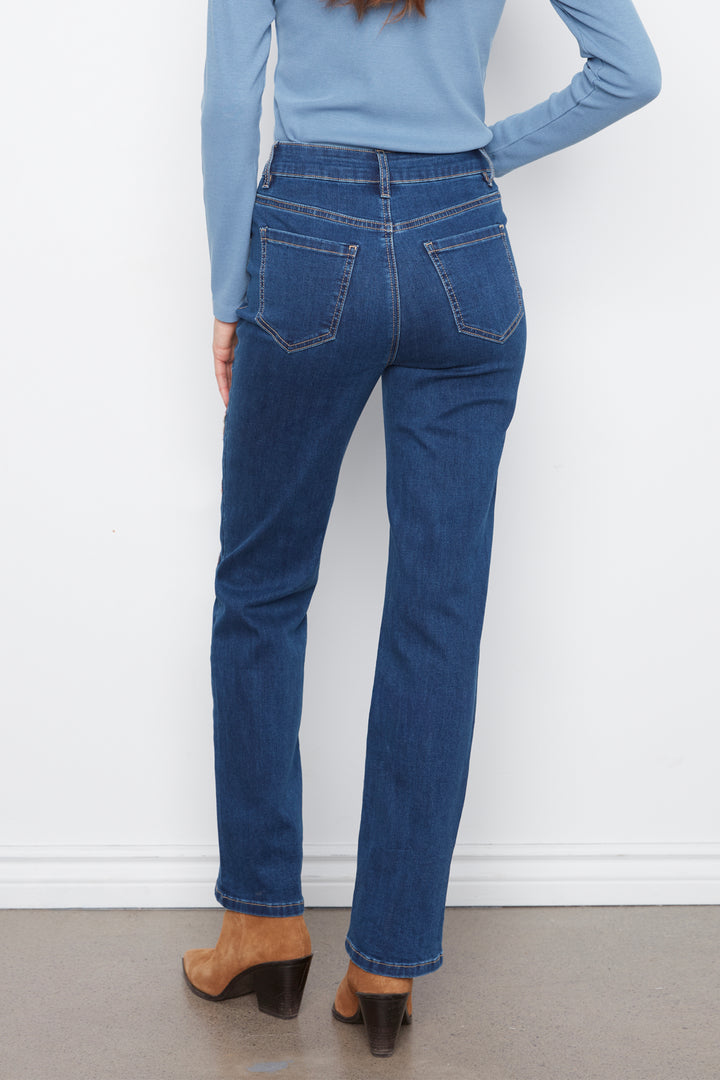 A person shown from the back wearing dark blue straight-leg jeans, a light blue long-sleeve top, and brown heeled sandals against a white wall and gray floor.