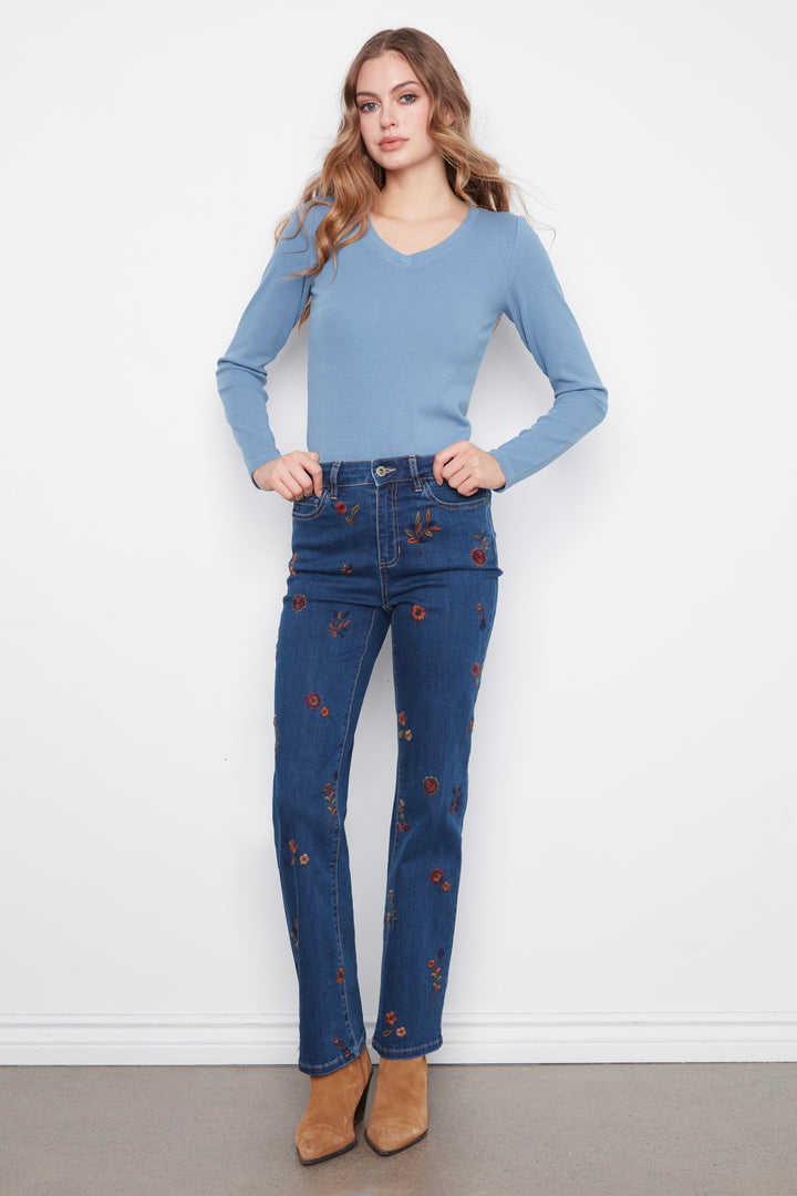 A woman modeling a pair of dark blue jeans with small red and yellow floral embroidery, paired with a light blue long-sleeve top and tan ankle boots, against a white wall.