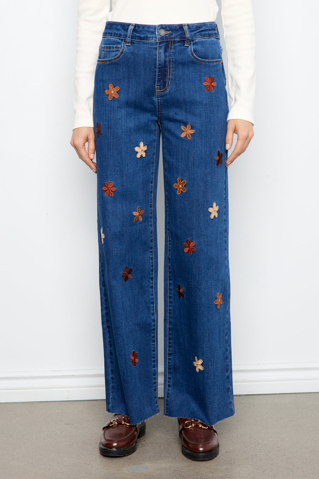 A person wearing blue wide-leg jeans with embroidered flower motifs in brown, orange, and cream, paired with a white long-sleeve top and brown loafers, standing against a white wall.