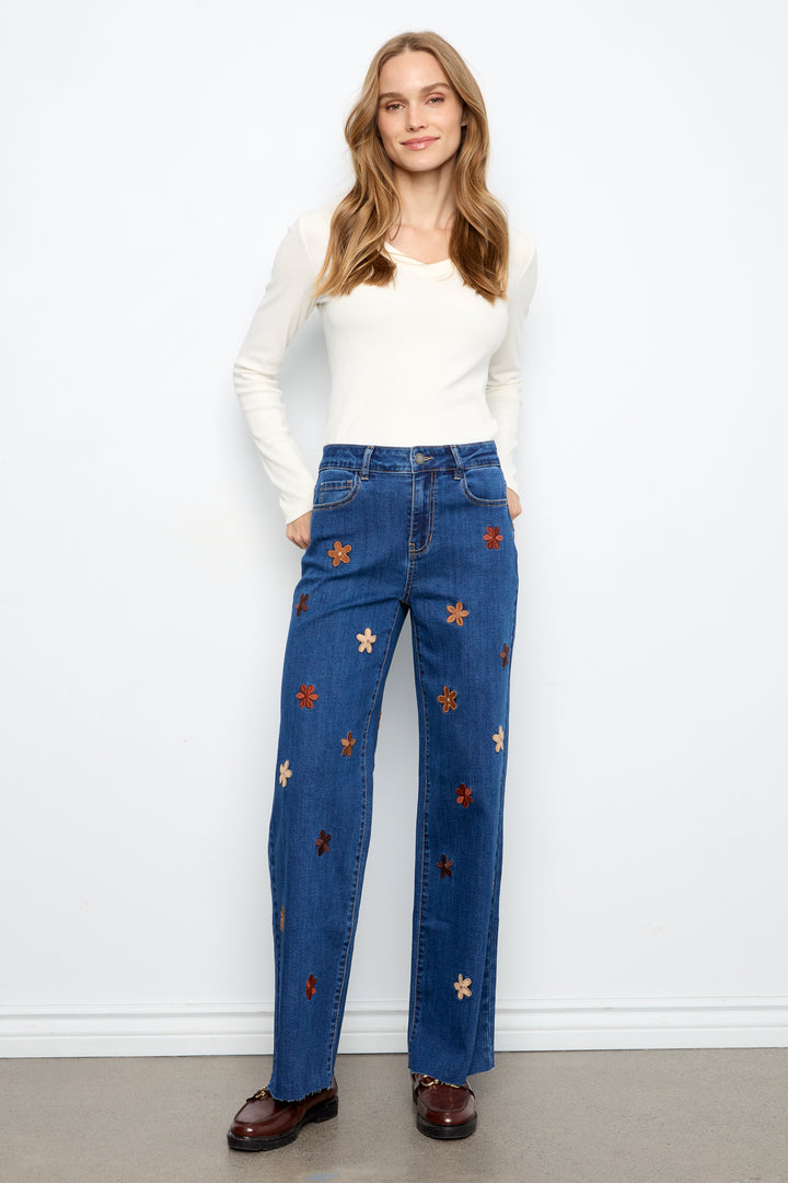 A woman modeling a pair of high-waisted, wide-leg blue jeans decorated with embroidered star patterns in gold, red, and white, paired with a white long-sleeve top and brown shoes.