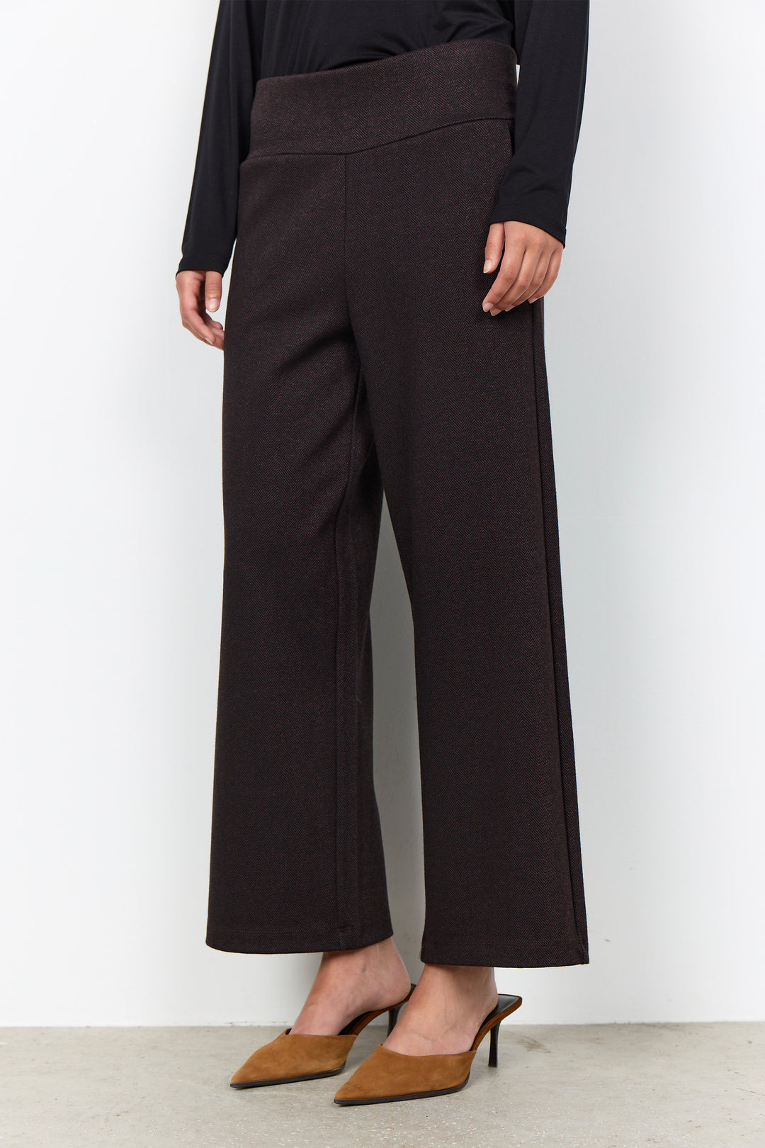A model wearing dark brown wide-leg cropped trousers with a matching elastic waistband, styled with a black long-sleeve top and brown pointed-toe mule heels against a plain white wall.