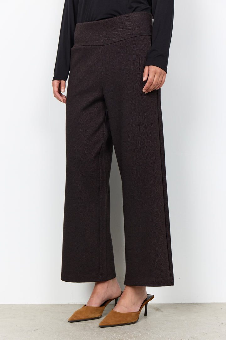A model wearing dark brown wide-leg cropped trousers with a matching elastic waistband, styled with a black long-sleeve top and brown pointed-toe mule heels against a plain white wall.