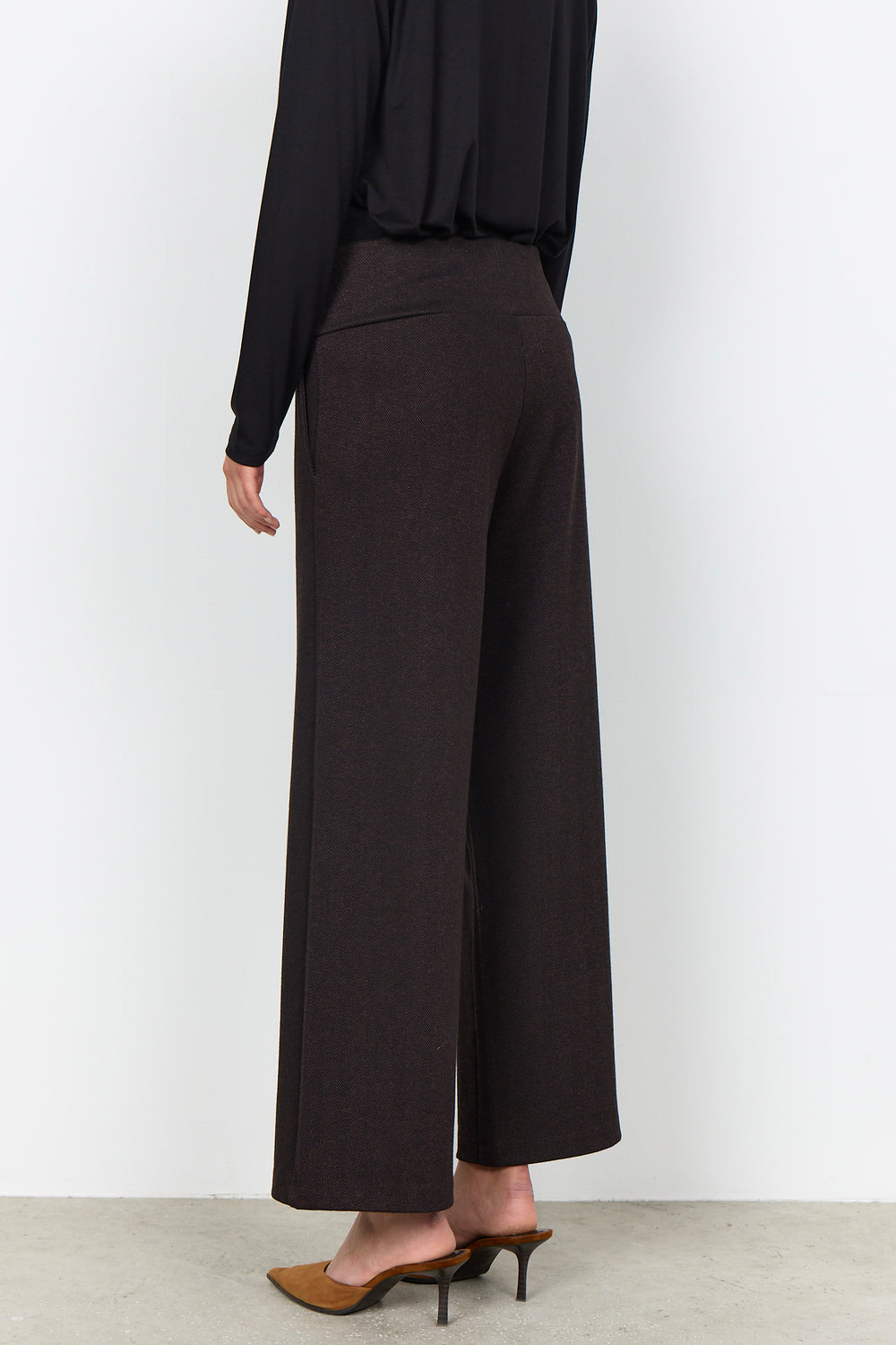 A model shown from the back wearing dark brown wide-leg cropped trousers, a black long-sleeve top, and brown pointed-toe mule heels against a plain white studio background.