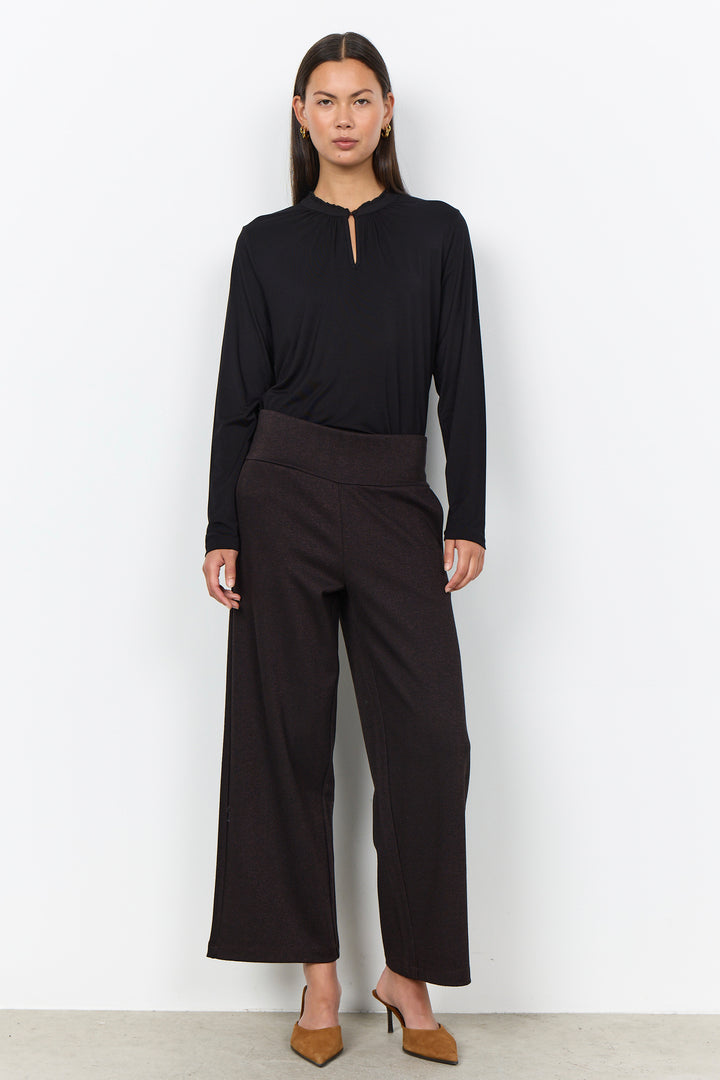 A woman modeling a black long-sleeve top with a keyhole neckline and black wide-leg cropped pants, paired with tan pointed-toe heels, against a plain white studio background.