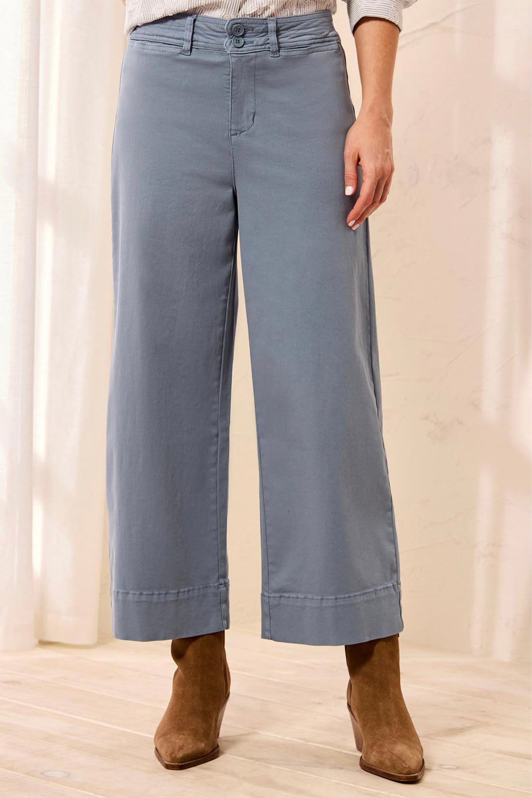 A person wearing wide-leg cropped pants in a dusty blue-gray color, paired with brown ankle boots, standing in a softly lit indoor setting with light curtains and a pale floor.
