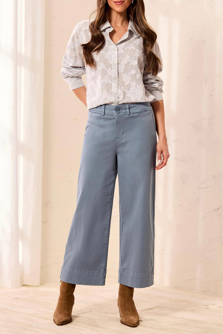 A woman models a pair of wide-leg cropped pants in a dusty slate blue color, paired with a white patterned button-up shirt and brown ankle boots, set against a warm beige interior background.