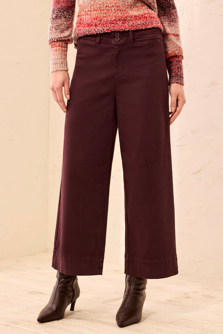 A woman wearing wide-leg cropped pants in a deep brown color, paired with brown ankle boots, standing in a softly lit indoor setting with light curtains and a pale floor.