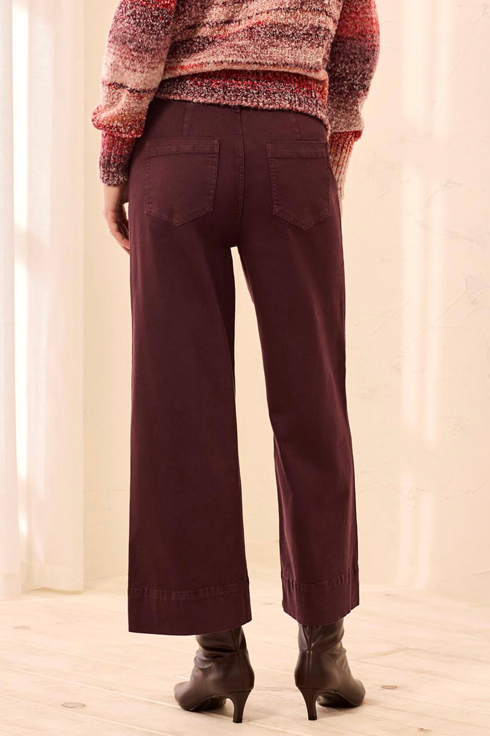 A woman shown from the back wearing wide-leg cropped pants in a deep brown color, paired with brown ankle boots, standing in a softly lit indoor setting with light curtains and a pale floor.