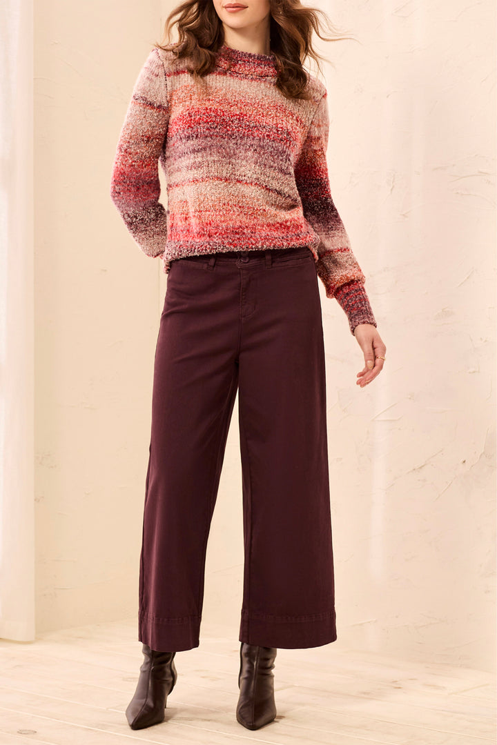 A woman wearing wide-leg cropped pants in a deep brown color, paired with brown ankle boots, standing in a softly lit indoor setting with light curtains and a pale floor.