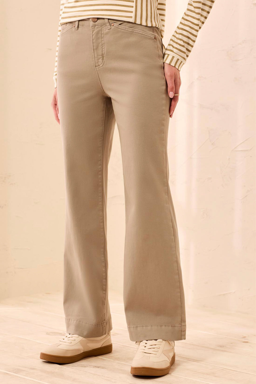 A person wearing beige straight-leg or bootcut pants, paired with a cream and tan striped long-sleeve top and cream sneakers, standing against a light beige textured wall.