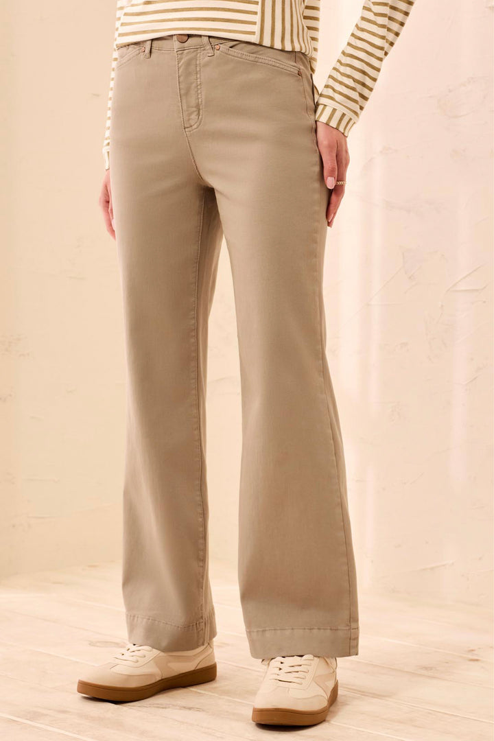 A person wearing beige straight-leg or bootcut pants, paired with a cream and tan striped long-sleeve top and cream sneakers, standing against a light beige textured wall.