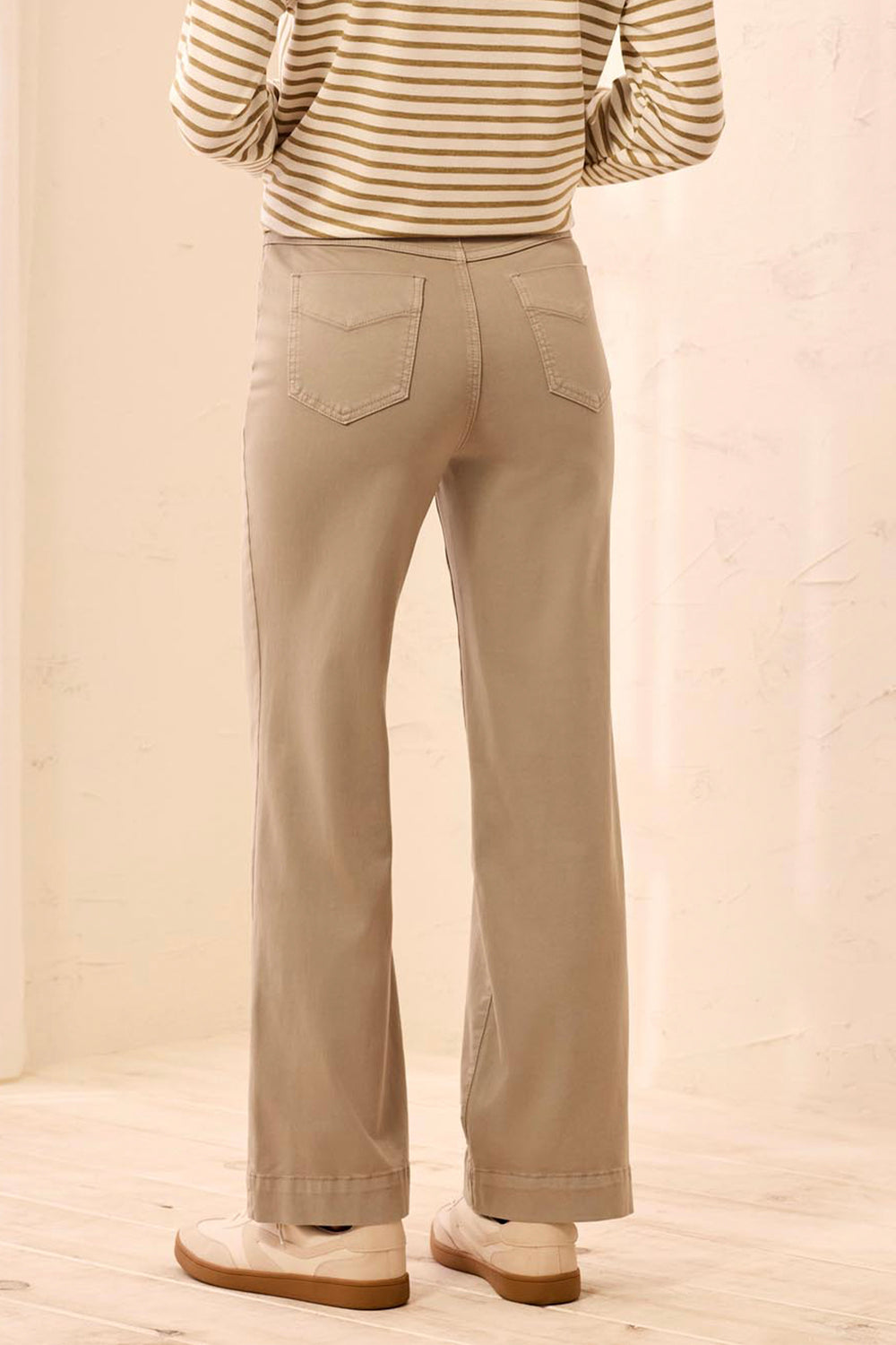 A person shown from the back wearing straight-leg beige pants, a beige and brown striped long-sleeve top, and light-colored shoes in a softly lit indoor setting.