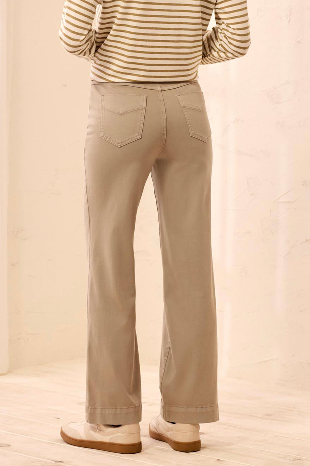A person shown from the back wearing straight-leg beige pants, a beige and brown striped long-sleeve top, and light-colored shoes in a softly lit indoor setting.