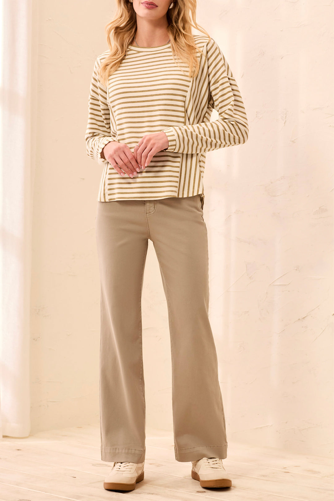 A woman modeling a beige and white striped long-sleeve top with beige wide-leg pants and white sneakers, standing in a bright indoor setting.