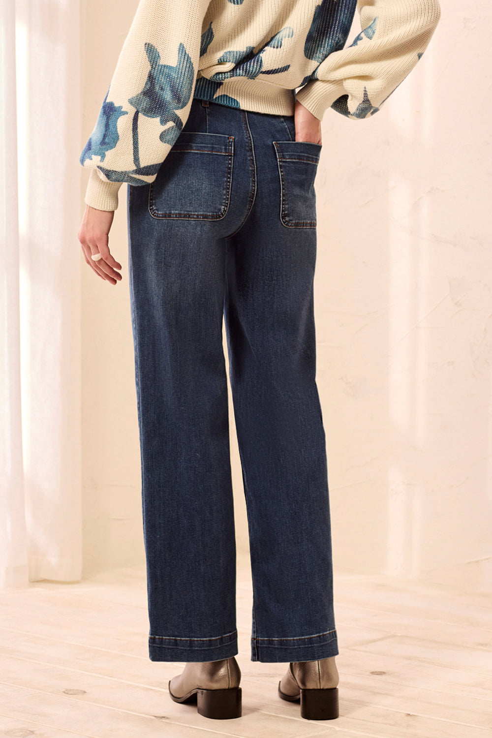 Rear view of a person wearing dark blue wide-leg jeans with a cream floral long-sleeve top and brown block-heel shoes in a softly lit indoor setting.