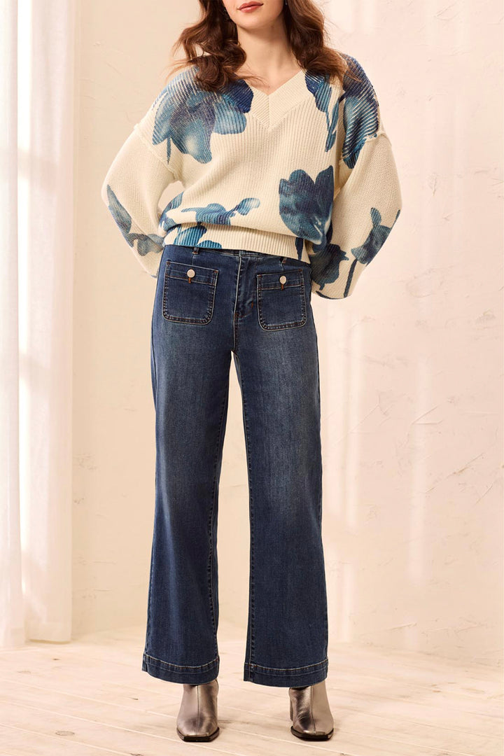 A model wearing a cream knit sweater with a blue floral pattern and dark blue wide-leg jeans, standing in a brightly lit indoor setting.