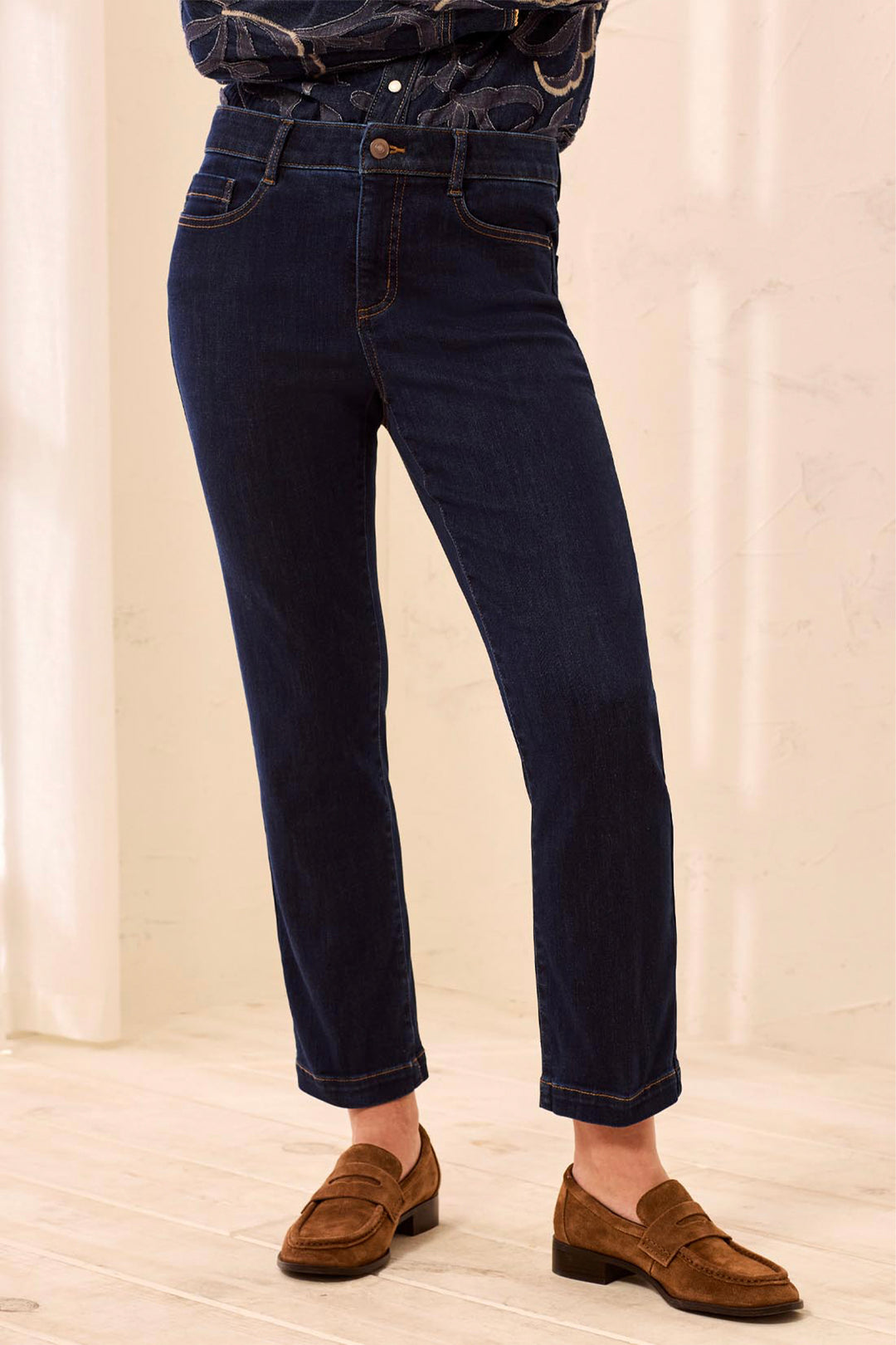 A person wearing dark blue slim-fit jeans with rolled cuffs, paired with brown suede penny loafers and a dark patterned top, standing in a bright indoor setting.