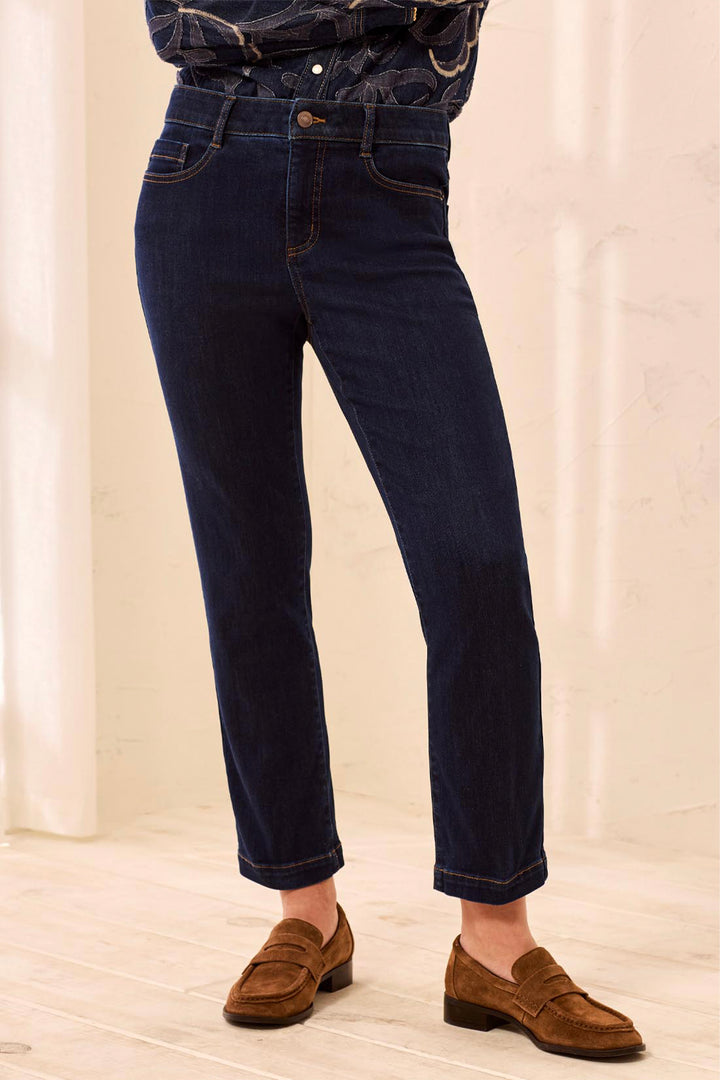 A person wearing dark blue slim-fit jeans with rolled cuffs, paired with brown suede penny loafers and a dark patterned top, standing in a bright indoor setting.