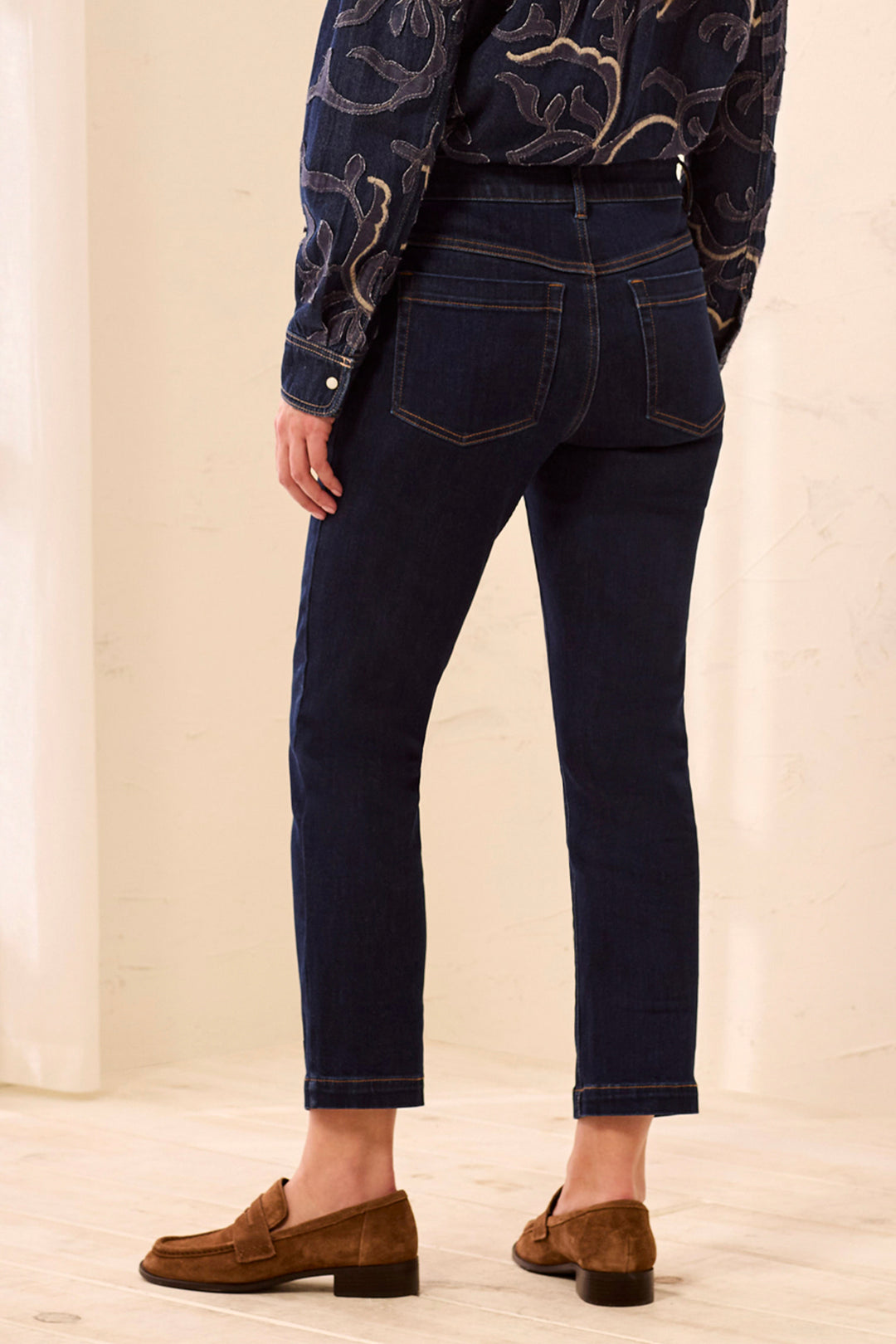 Rear view of a person wearing dark blue slim-fit jeans with contrast stitching, paired with a dark patterned blouse and brown suede loafers, standing against a light cream background.