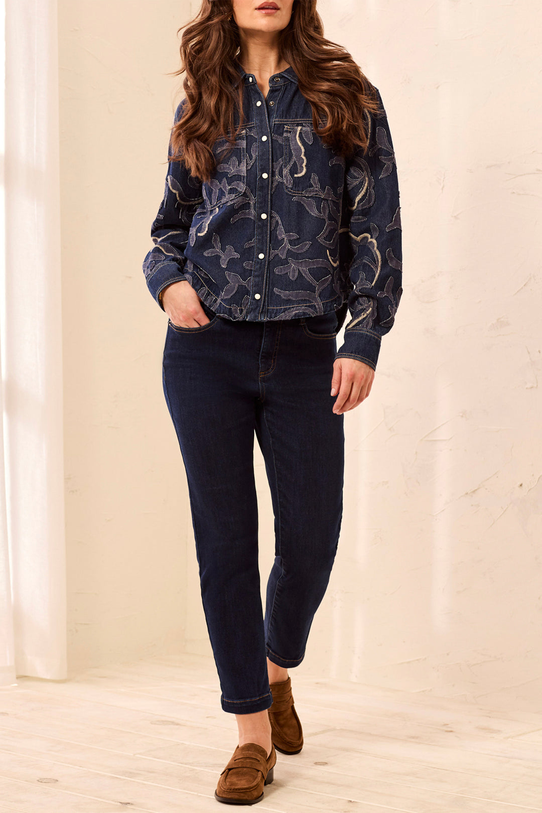 A woman modeling a dark navy blue patterned button-up shirt jacket with dark blue jeans and brown suede loafers, standing in a brightly lit indoor setting with a cream-colored wall and light wood floor.