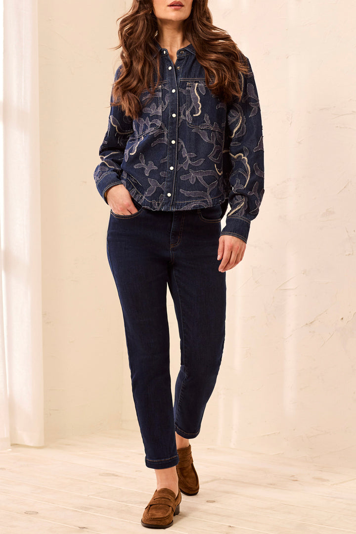 A woman modeling a dark navy blue patterned button-up shirt jacket with dark blue jeans and brown suede loafers, standing in a brightly lit indoor setting with a cream-colored wall and light wood floor.