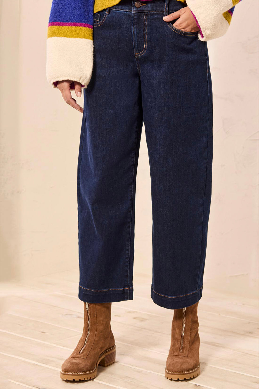 A person wearing dark blue wide-leg cropped jeans with rolled cuffs, paired with brown ankle boots and a multicolored knit sweater, standing against a light beige wall and wood floor.