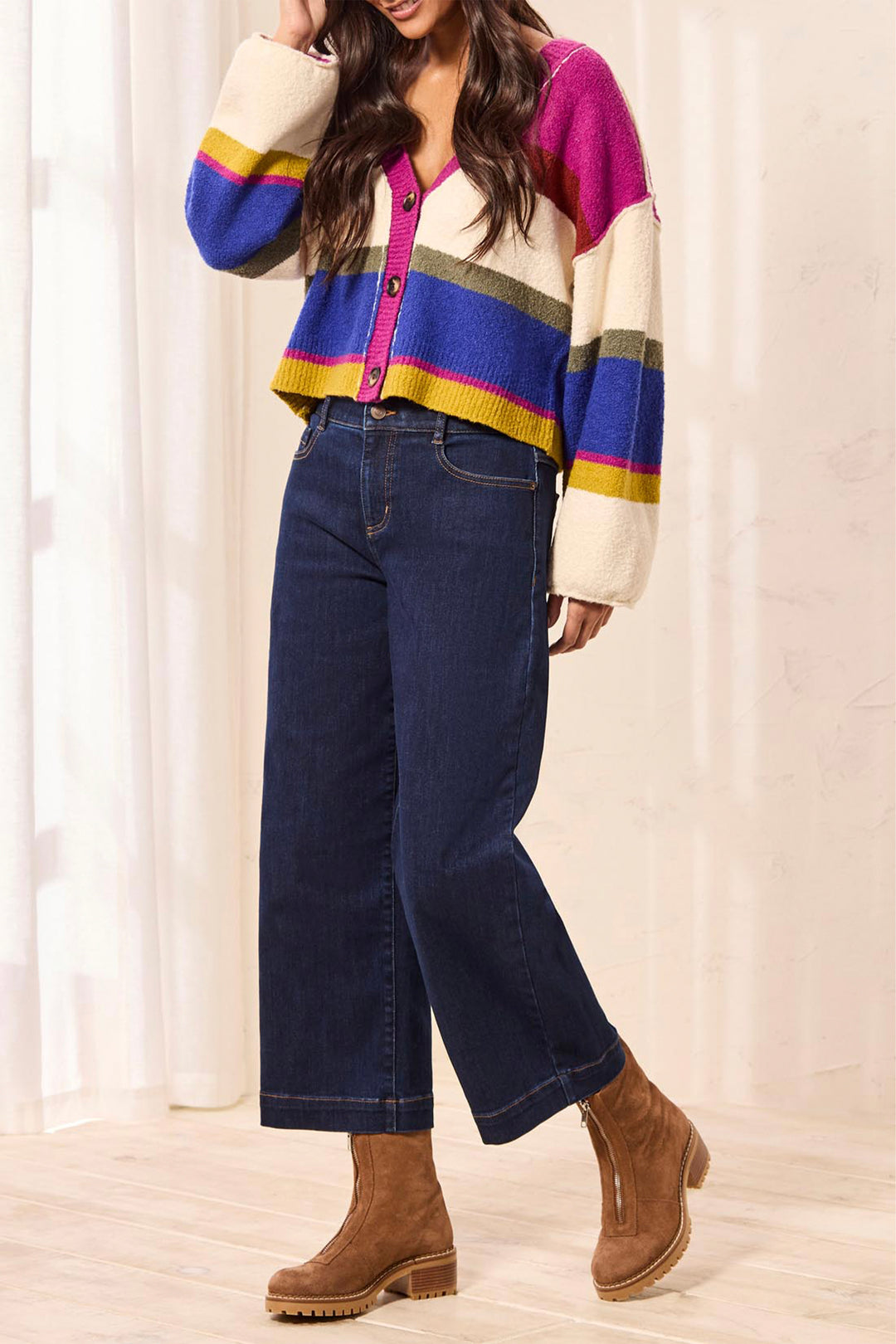 Person wearing a colorful striped sweater and blue jeans with brown boots against a white curtain background