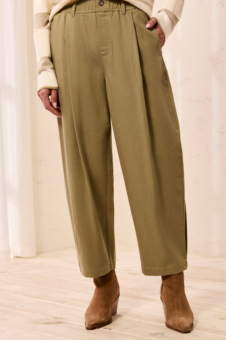 A person wearing wide-leg pleated trousers in a muted olive-tan color, paired with a cream top and brown ankle boots, standing against a light neutral background.