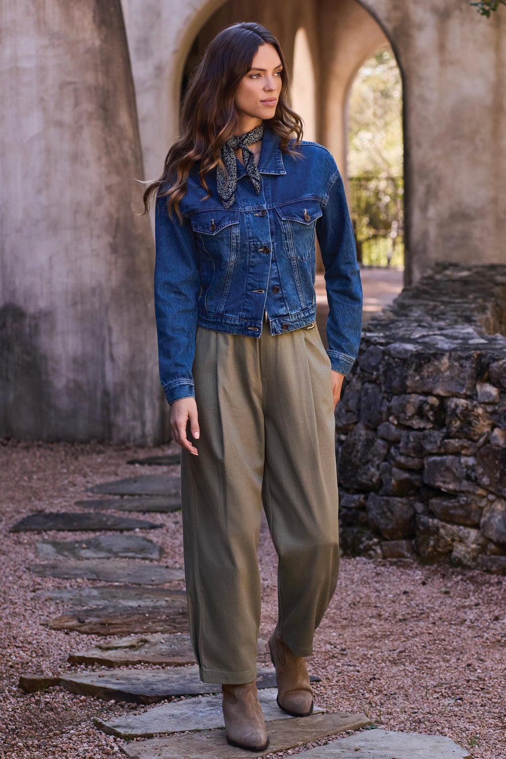 A woman stands outdoors in a stone archway setting wearing a cropped blue denim jacket, a patterned neck scarf, loose olive green wide-leg pants, and brown ankle boots.