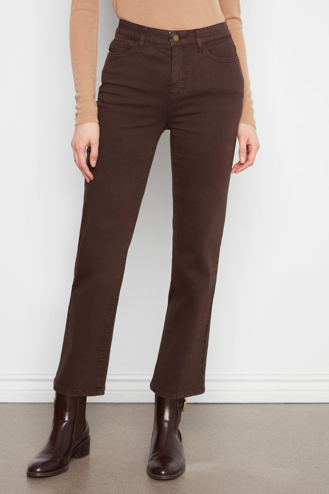 A model wearing dark brown straight-leg jeans, a beige long-sleeve top, and dark brown leather ankle boots, standing against a white wall.