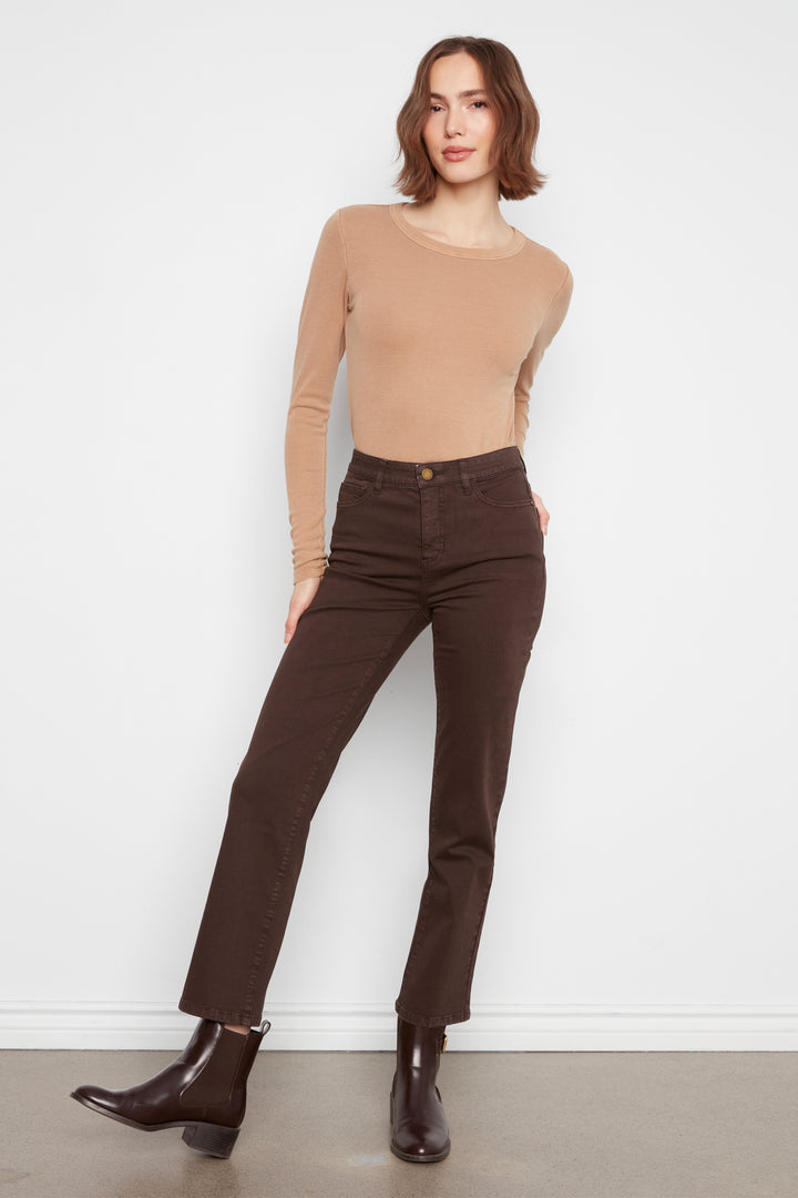 A woman modeling a beige long-sleeve knit top and dark brown straight-leg jeans with dark brown ankle boots, standing against a white wall.