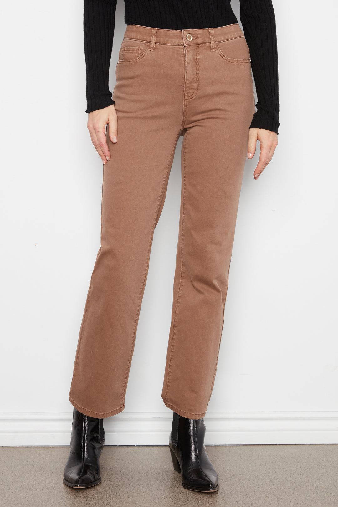 A person wearing straight-leg brown jeans with a black long-sleeve top and black ankle boots, standing against a white wall.