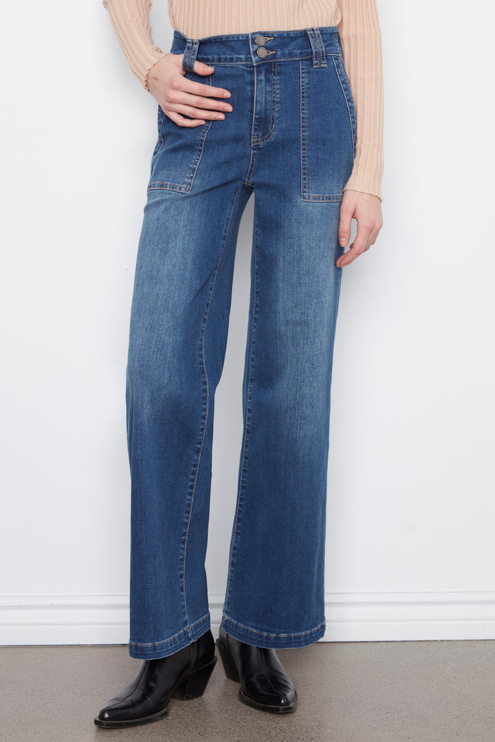A woman modeling a beige ribbed mock-neck long-sleeve top and high-waisted, wide-leg blue denim jeans with black leather boots, standing against a white wall.