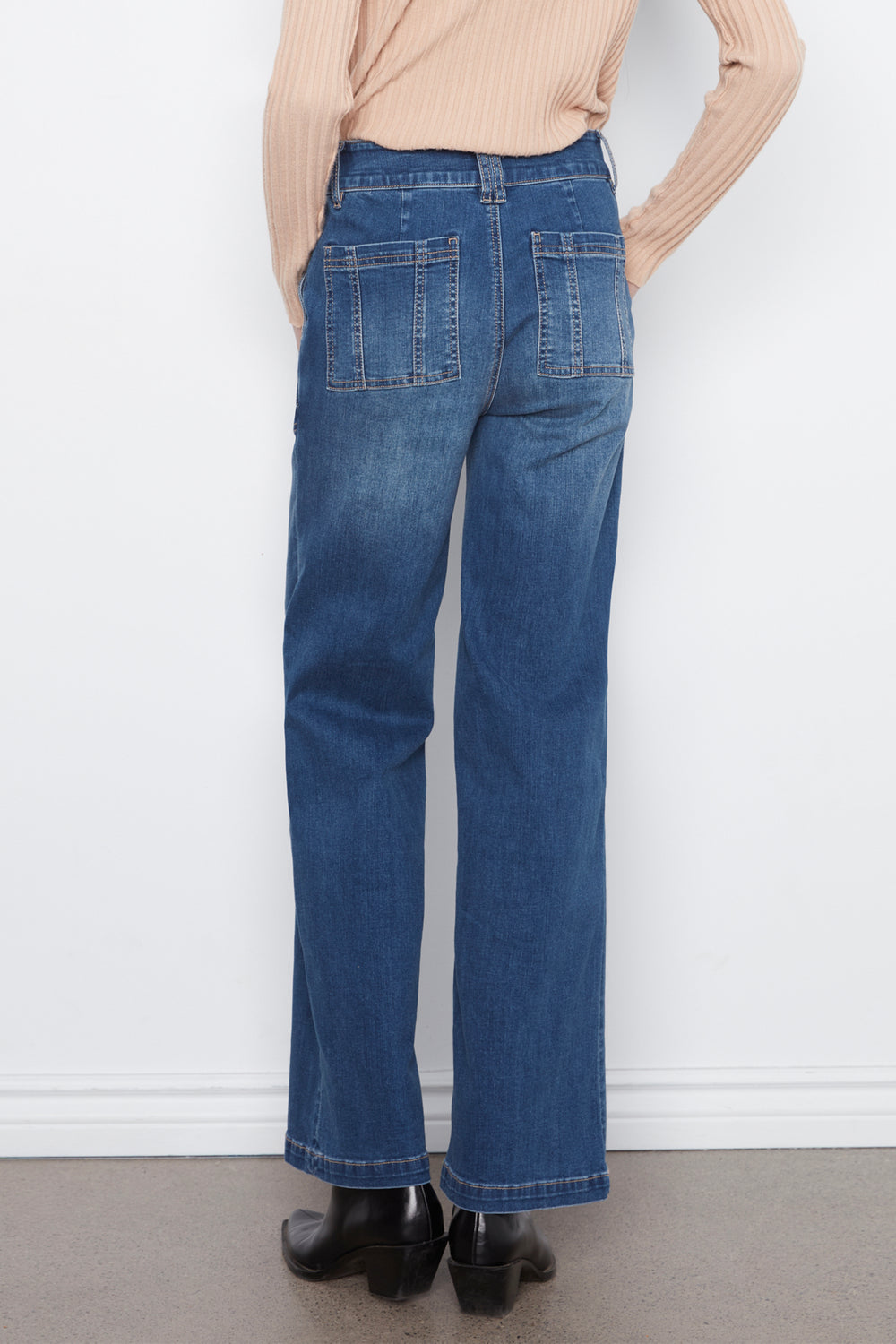 A person shown from the back wearing blue wide-leg jeans, a beige ribbed long-sleeve top, and black ankle boots against a plain white wall.