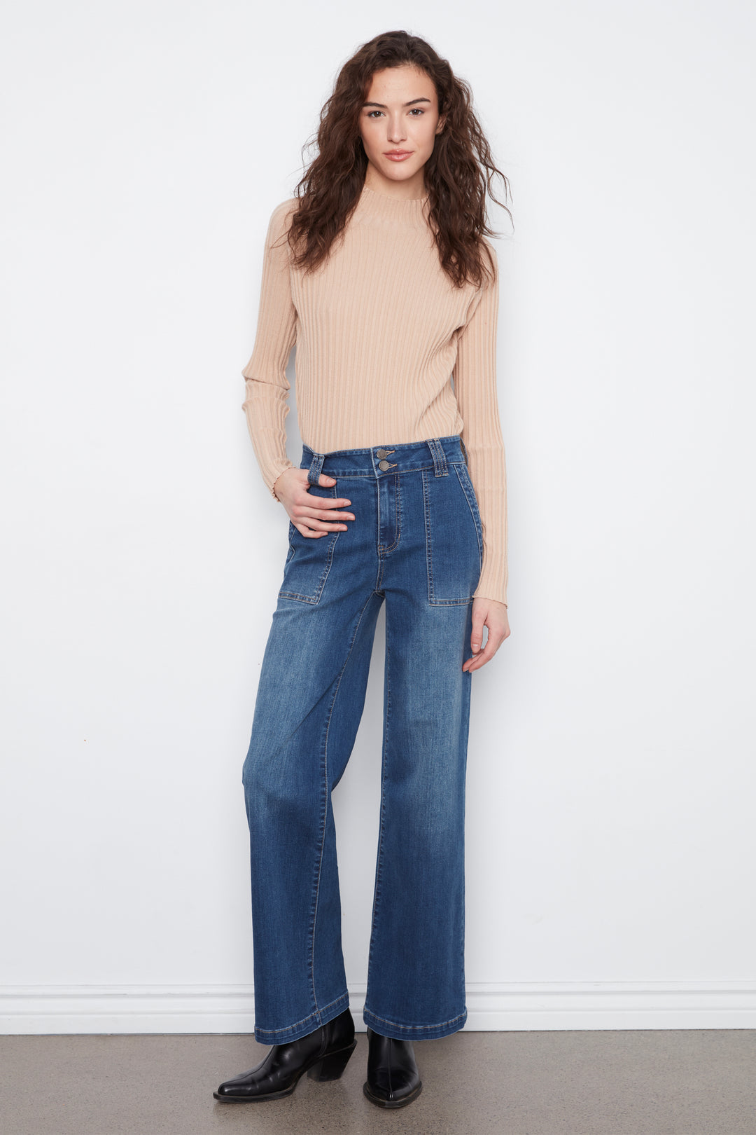 A woman modeling a beige ribbed mock-neck long-sleeve top and high-waisted, wide-leg blue denim jeans with black leather boots, standing against a white wall.