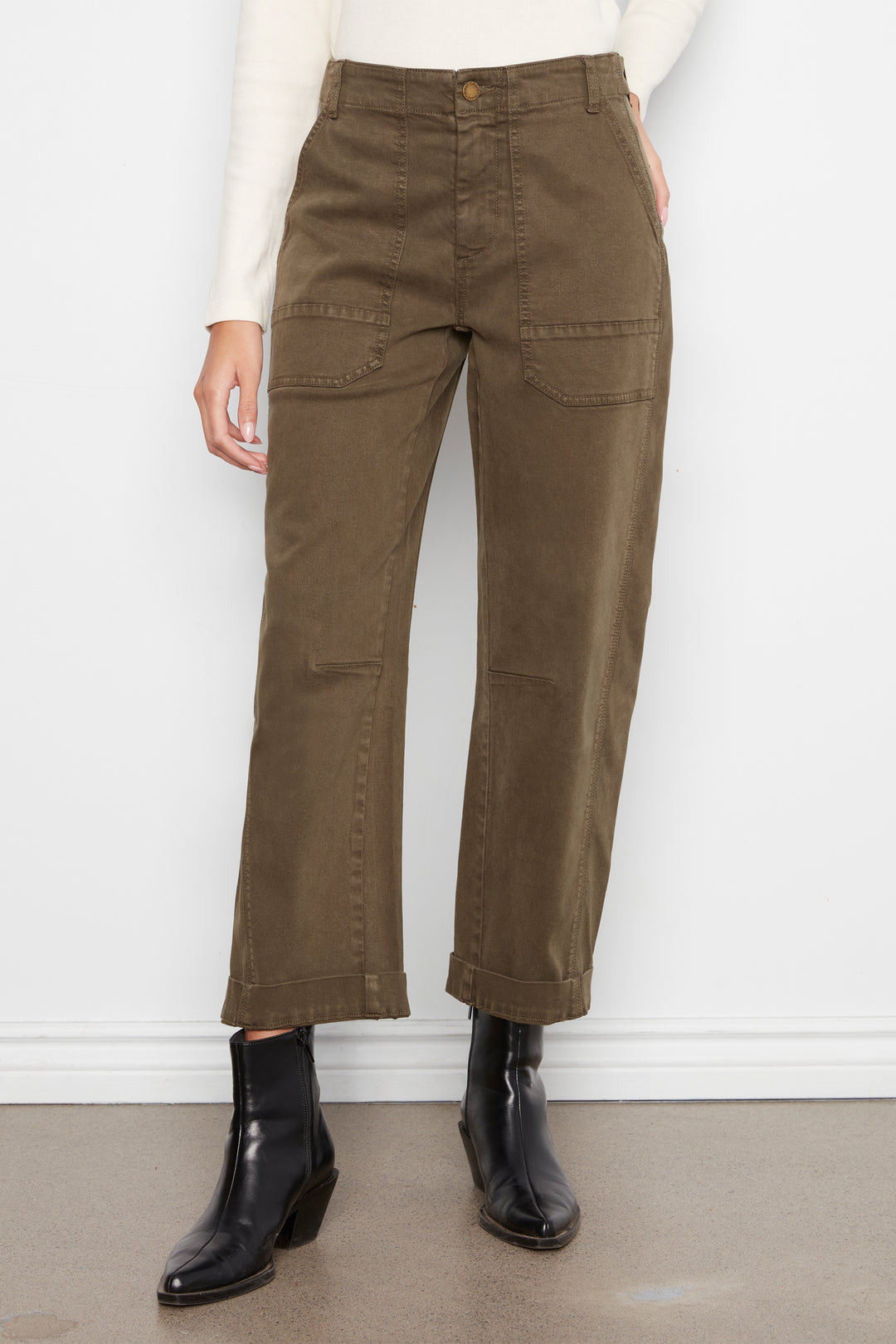 A person wearing olive brown cropped cargo pants with a cream long-sleeve top and black ankle boots, standing against a white wall.