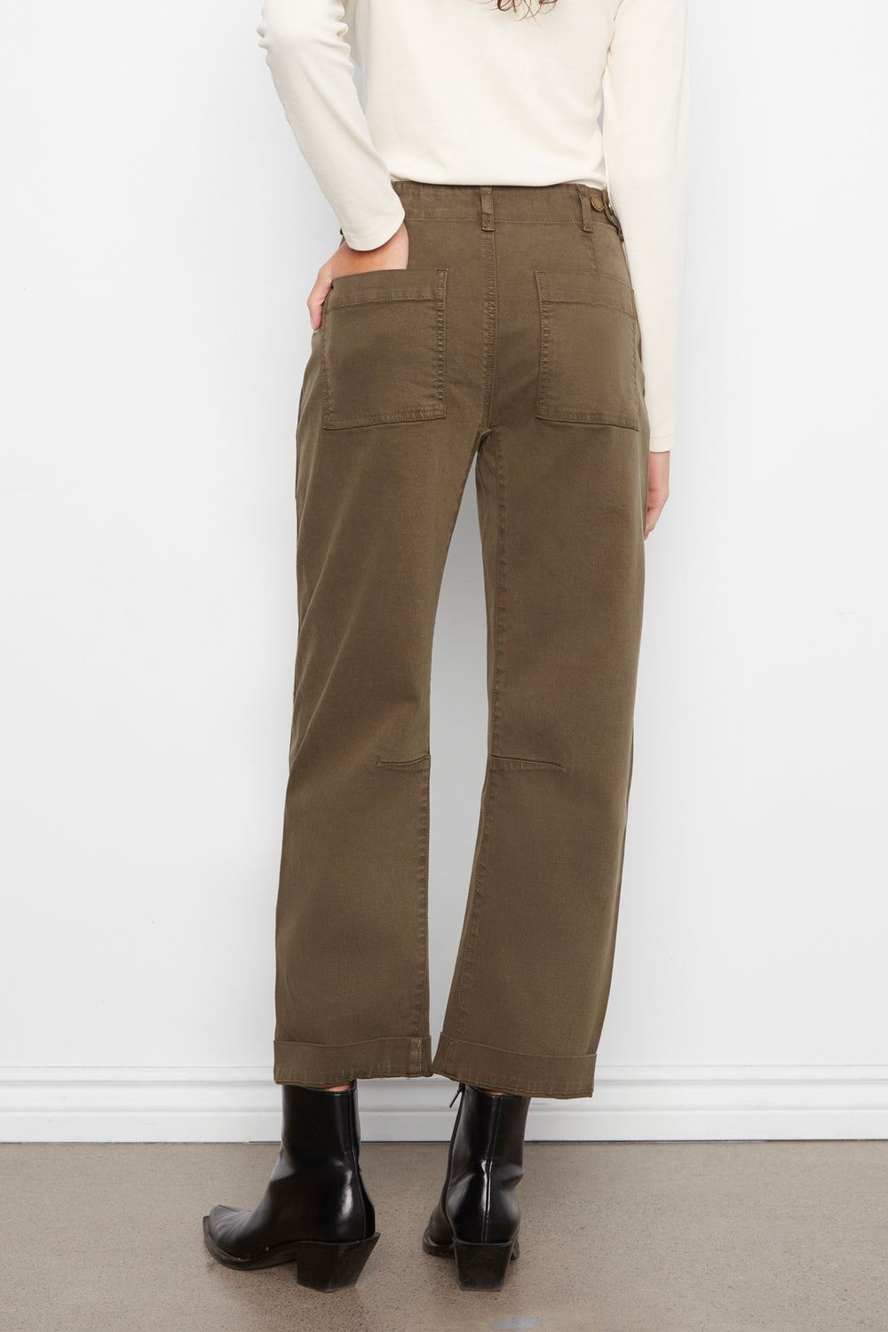 A person shown from the back wearing olive brown wide-leg cropped pants, a cream long-sleeve top, and black ankle boots against a white wall.