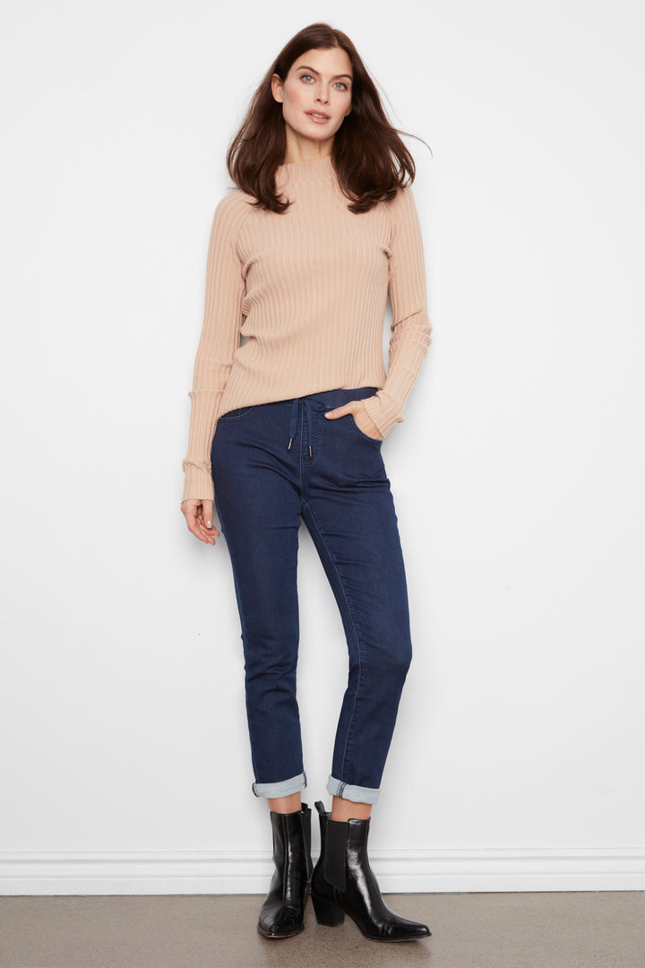 A person wearing dark blue slim-fit jeans with rolled cuffs, paired with a beige ribbed sweater and black ankle boots, standing against a white wall.