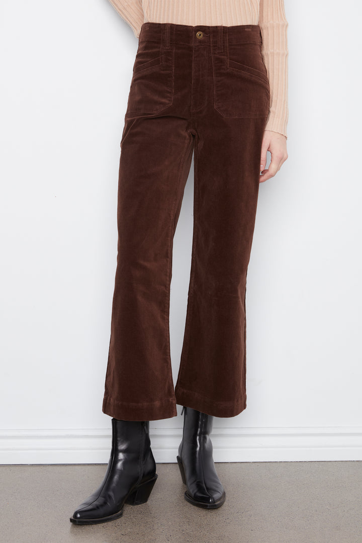 A person wearing dark brown corduroy cropped flare pants, a beige ribbed knit top, and black ankle boots, standing against a white wall.