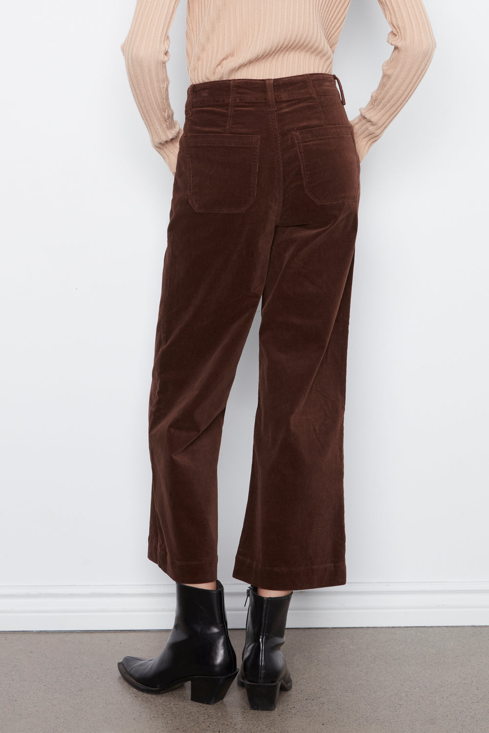 A person shown from the back wearing dark brown wide-leg corduroy pants, a beige ribbed sweater, and black ankle boots against a white wall.