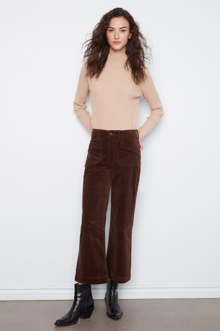 A woman modeling a beige ribbed knit mock-neck top and dark brown wide-leg corduroy pants, paired with black ankle boots, standing against a plain white wall.