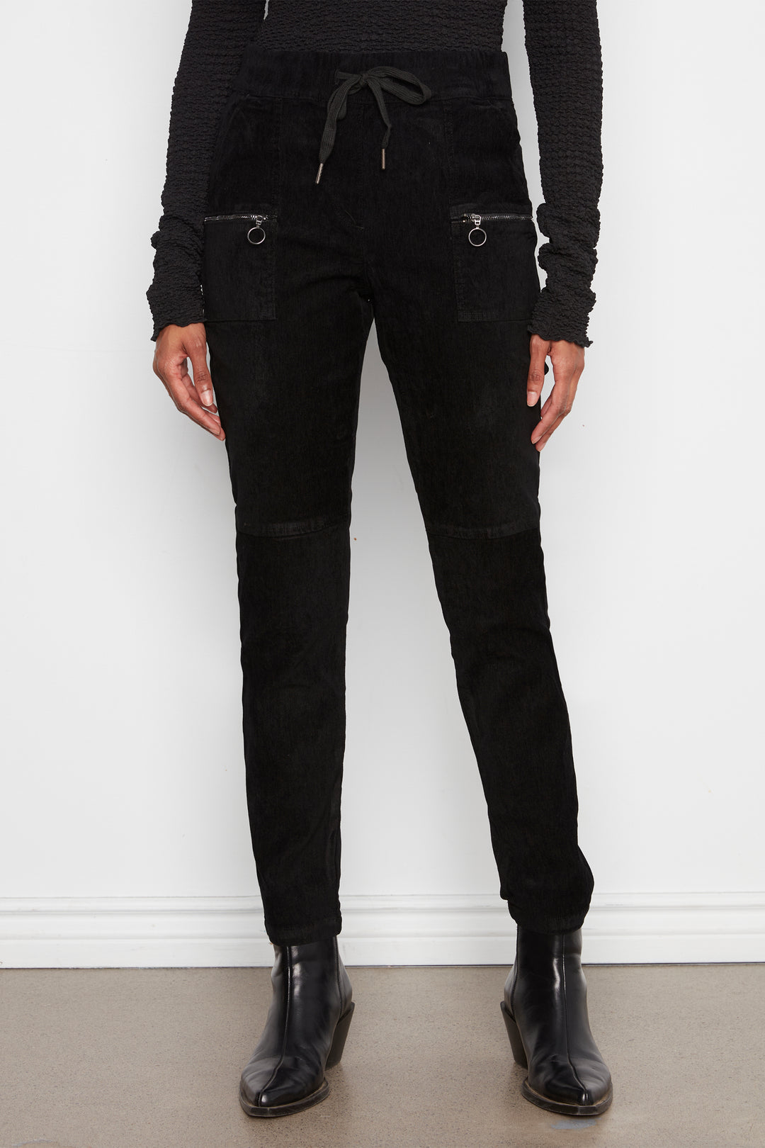 A model wearing black corduroy pants with zippered pockets and a drawstring waist, paired with a black ribbed long-sleeve top and black ankle boots, standing against a white wall.
