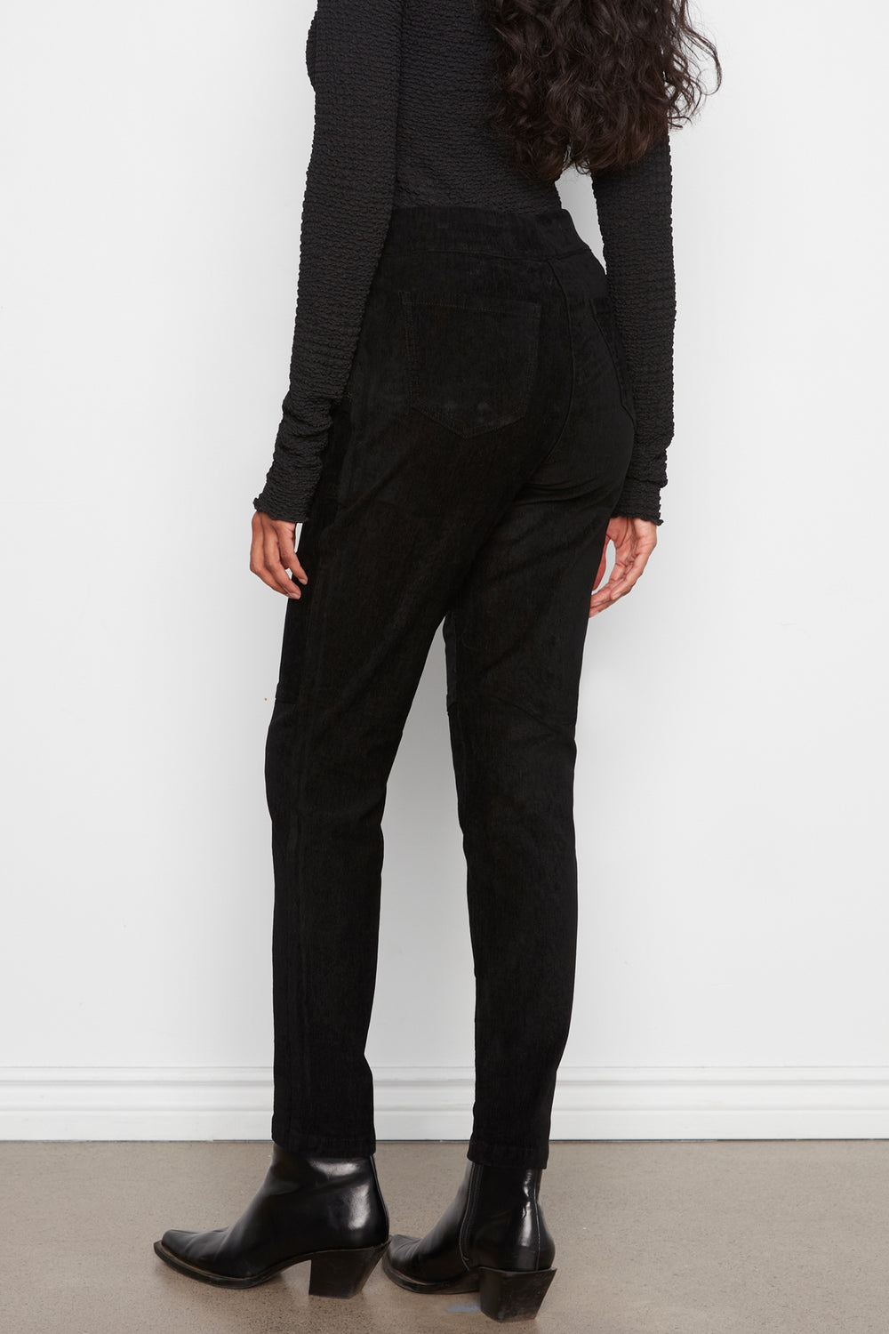 A model shown from the back wearing black high-waisted trousers, a black ribbed long-sleeve top, and black leather ankle boots against a white wall and beige floor.